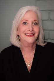 A smiling woman with silver hair and a black top poses against a light background, looking warm and friendly.