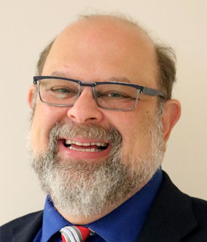 Eric Jacobson, a white man, wears glasses and smiles at the camera. He is bald-headed, with a graying beard and wears a dark suit. Eric Jacobson, a white man, wears glasses and smiles at the camera. He is bald-headed, with a graying beard and wears a dark suit.
